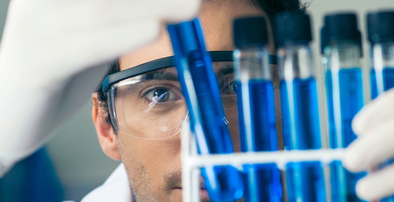 A man wearing goggles stares into a tray of test tubes.